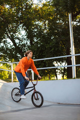 Young guy in orange pullover and jeans riding bicycle at skatepark