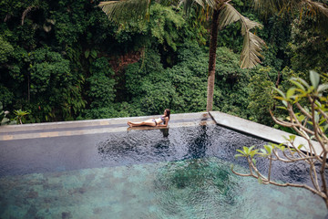 Slim sexy brunette woman in swimsuit relaxing on edge tropical infinity pool in jungle. Palms around and crystal clean water. Luxury resort on Bali island.