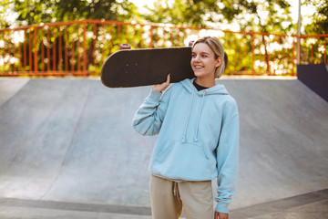 Pretty smiling girl in hoodie holding skateboard on shoulder while happily looking aside at...