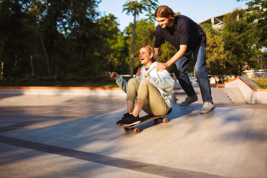 Pretty cheerful girl sitting on skateboard and riding with young guy near while joyfully spending time together at skatepark