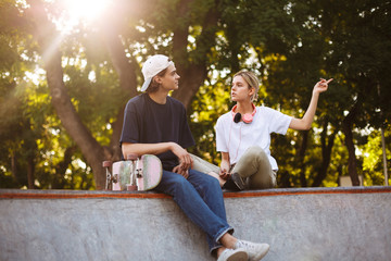 Beautiful girl with headphones and young guy with skateboard thoughtfully discussing something together while spending time at modern skatepark