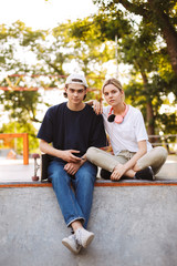 Young guy with cellphone and pretty girl with headphones dreamily looking in camera while spending time together at skatepark