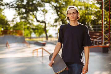Young serious guy in black T-shirt and white cap thoughtfully looking in camera while holding skateboard in hand with modern skatepark on background