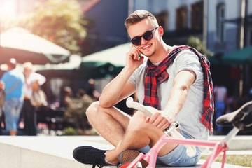 Fototapeta na wymiar Handsome young hipster man in sunglasses sitting near the bicycle and speaking on the smartphone in the sunny street