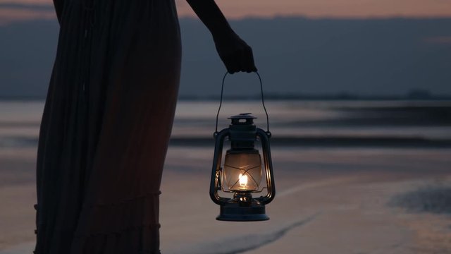 Girl in dress holding old kerosene lantern in her hand at beach after sunset at night