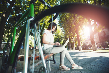 Cute romantic girl with green bicycle sitting on beanch and working with laptop in the sunny summer park