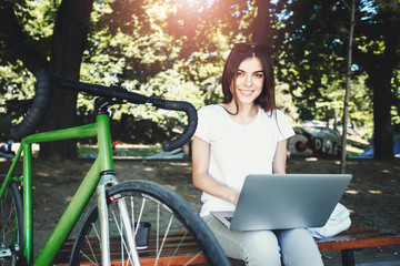 Naklejka na ściany i meble Cute romantic girl with green bicycle sitting on beanch and working with laptop in the sunny summer park, she smiling