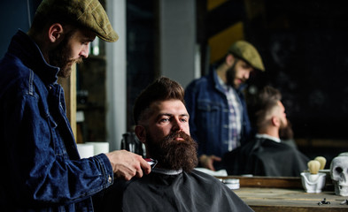 Hipster with beard covered with cape trimming by professional barber in stylish barbershop. Barber busy with grooming beard of hipster client, mirror reflexion on background. Grooming concept