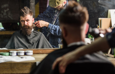 Barber with clipper trimming hair on nape of client. Reflexion of barber with hair clipper works on haircut of guy, barbershop background. Hipster hairstyle concept. Hipster client getting haircut