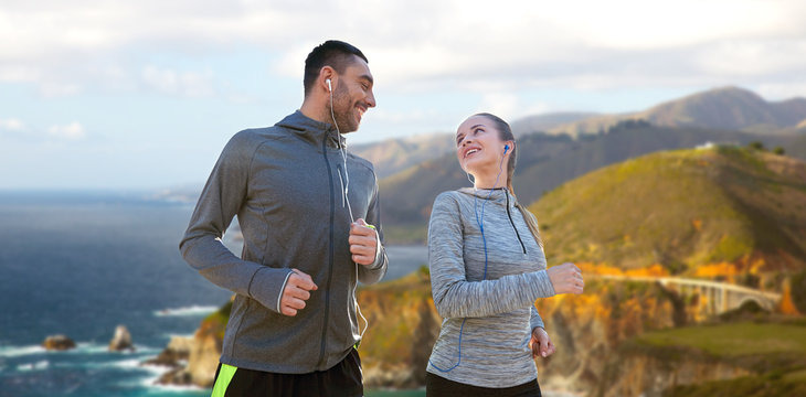fitness, sport and technology concept - happy couple running and listening to music in earphones over bixby creek bridge on big sur coast of california background