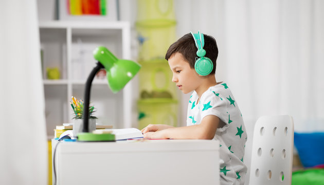 education, childhood and school concept - student boy in headphones reading book at home