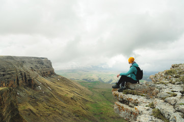 Obraz premium Young woman with a backpack pensively sitting on the edge of a rock and looking at the sky with clouds