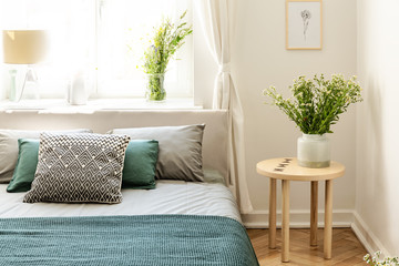 Flowers on wooden table next to bed with pillows and green sheets in bedroom interior. Real photo