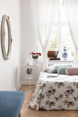 White bedside table with fresh red roses, candles and coffee cup in real photo of bright bedroom interior with window with curtains