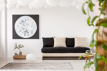 Pillows on black sofa in white living room interior with moon poster and grey carpet. Real photo
