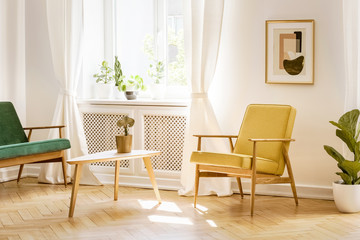 Yellow, retro armchair and a green sofa by a big, sunny window in a stylish, white real living room interior with a poster on the wall