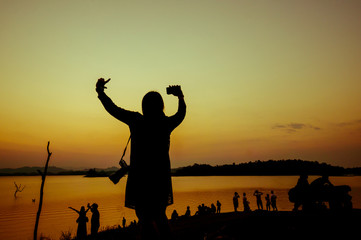 The silhouette of a woman raise her hands up among the crowd near the water in the sunset