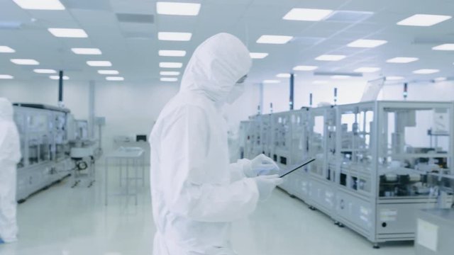 Scientist Using Digital Tablet Computer and wearing Protective Suit walks through Manufacturing Laboratory with Modern Industrial Machinery.