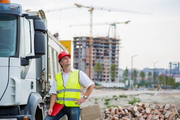 Truck driver in front of the truck on a construction site with buildings and cranes in the...