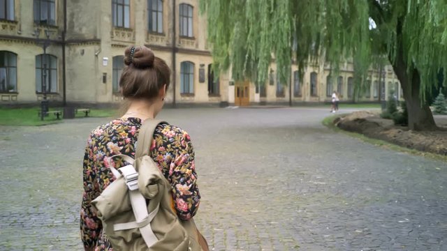 Young cheerful female college student with backpack going to college, walking on street near university, beautiful and charming