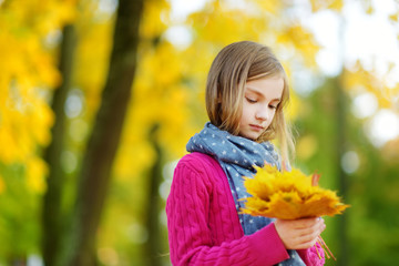 Cute little girl having fun on beautiful autumn day. Happy child playing in autumn park. Kid gathering yellow fall foliage.