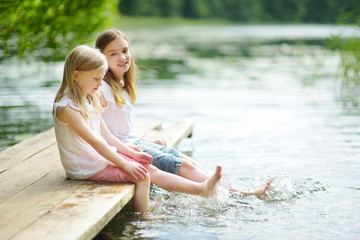 Two cute little girls sitting on a wooden platform by the river or lake dipping their feet in the water on warm summer day