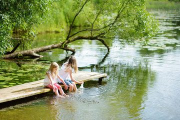 Two cute little girls sitting on a wooden platform by the river or lake dipping their feet in the...