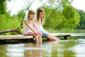 Two cute little girls sitting on a wooden platform by the river or lake dipping their feet in the...