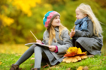 Cute little girls sketching outside on beautiful autumn day. Happy children playing in autumn park. Kids drawing with colourful pencils.