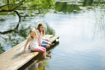 Two cute little girls sitting on a wooden platform by the river or lake dipping their feet in the...