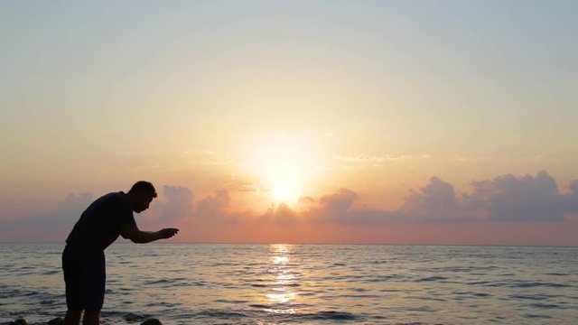 Black silhouette of adult tall man taking pictures of beautiful scenic seascape and selfie during sunrise on early morning. Real time full hd video footage.