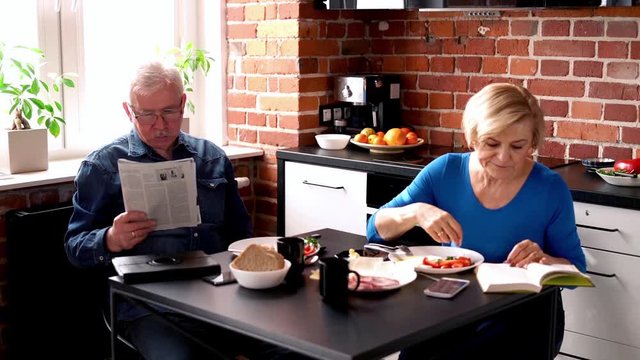 Senior couple sitting with book and newspaper in the kitchen during breakfast 
