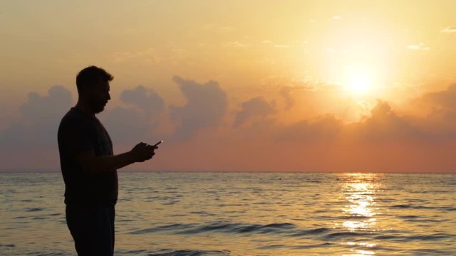 Black silhouette of adult tall man taking pictures of beautiful scenic seascape and selfie during sunrise on early morning. Real time full hd video footage.