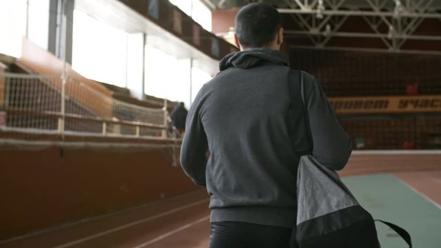 Tracking with rear view of unrecognizable sportsman with gym bag walking along track in indoor stadium and taking off hood