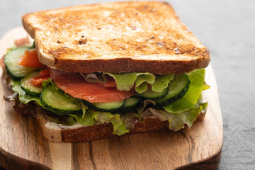 Sandwich with white bread toasts, red fish salmon, fresh green leaves of salad and sliced cucumber on a wooden board.