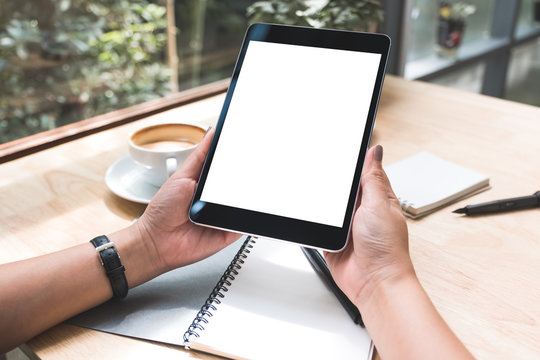Mockup image of a woman's hands holding black tablet pc with white blank screen with notebook and coffee cup on table