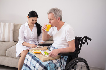 Beautiful asian nurse with her hair in sleek ponytail sitting on couch next to male patient with gray hair in wheelchair eating lunch.
