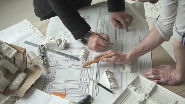 Hands of two male designers working on molding blueprint on the table with white plaster samples in their studio, using pensils in their hands.