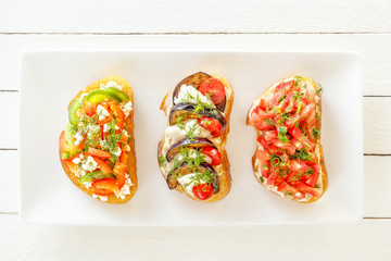 Bruschetta with fresh tomatoes and cheese, eggplant and mozzarella, sweet pepper and goat cheese on a white plate. Top view