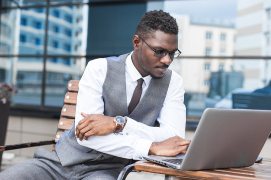 african businessman in suit and glasses is sitting at a table with a Laptop, a cup of coffee against a background of glass buildings