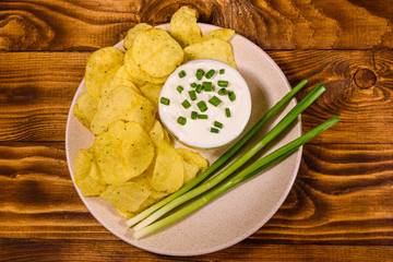 Ceramic plate with potato chips and glass bowl with sour cream on wooden table. Top view