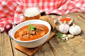 Close-up of pork goulash with pieces of meat in a bowl, garlic, pepper, onion, glass with cold beer and red checkered tablecloth in the background