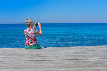 Kobieta z aparatem fotograficznym, siedząca na drewnianym pomoście nad morzem
