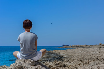 Fototapeta na wymiar Dziewczyna siedząca na skale nad morzem, patrząca na wrak statku MV Demetrios II, Paphos, Cypr