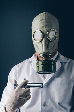 Creepy medical experiment concept, a scary doctor in gas mask with a big stainless steel syringe on dark background
