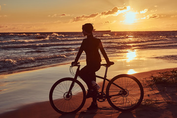 Fototapeta premium Female cyclist standing with her bicycle and enjoying the sunset on the sea coast.