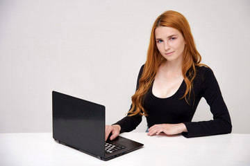 portrait of a beautiful girl with red hair on a white background sitting at the table and working behind a laptop.