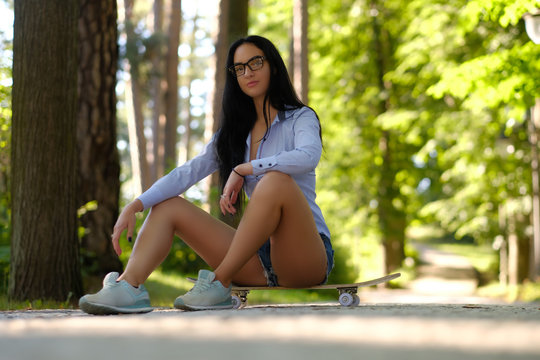 Gorgeous sensual brunette girl in glasses wearing a shirt and shorts sitting on a skateboard in a park.