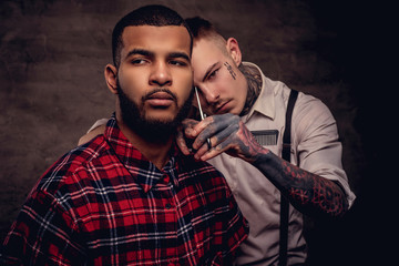 Old-fashioned professional tattooed hairdresser does a haircut to an African American client. Isolated on dark textured background.