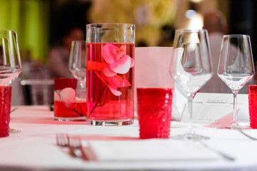 Restaurant table set with white tablecloth and red details, such as glasses and a vase with red water and an orchid inside